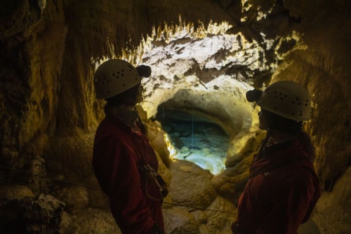 Two explorers wearing helmets and red suits stand inside a glowing Banff cave, admiring a crystal-blue underground pool.